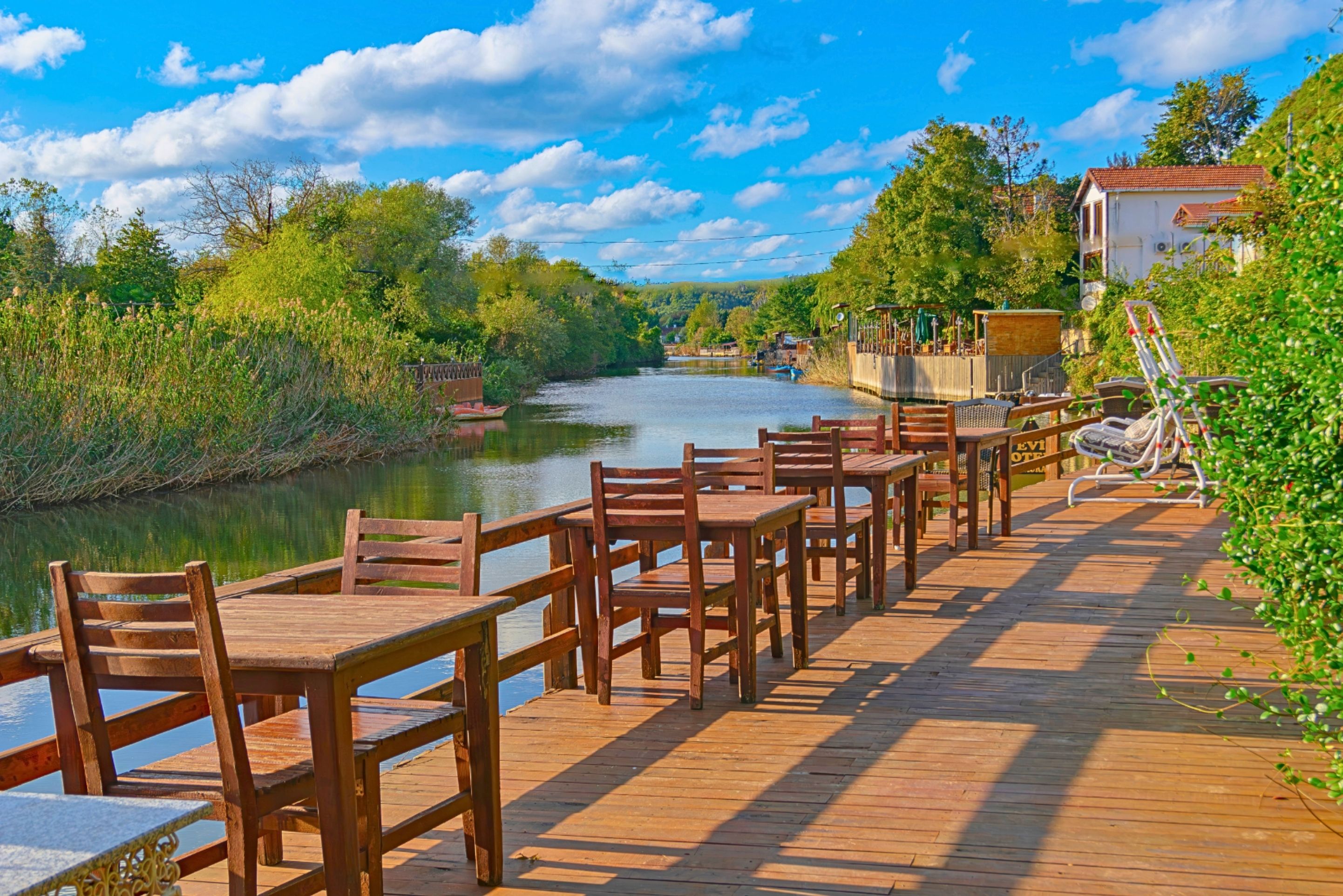 Une terrasse en bois, agrémentée de tables et de chaises, qui offre une vue sur une rivière et est entourée de verdure et de bâtiments.