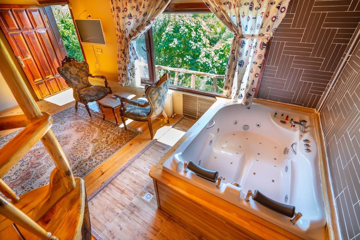 Quarto com piso de madeira, uma banheira grande, duas cadeiras, uma mesa pequena e uma janela com cortinas.