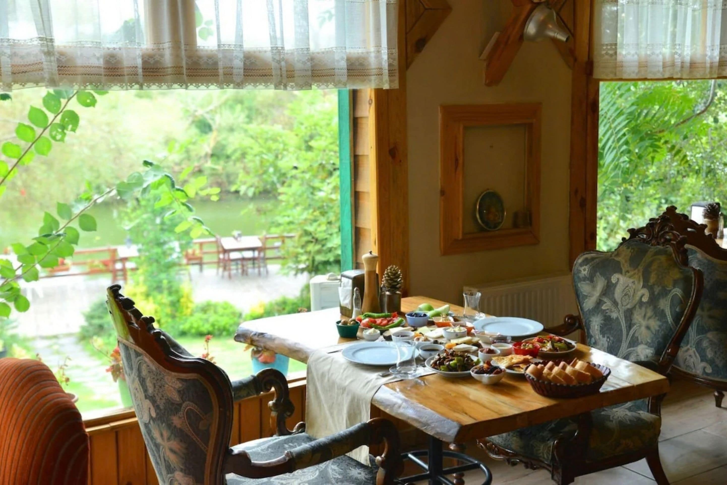 Un comedor con una mesa dispuesta para una comida, sillas y vistas de un jardín a través de una ventana.