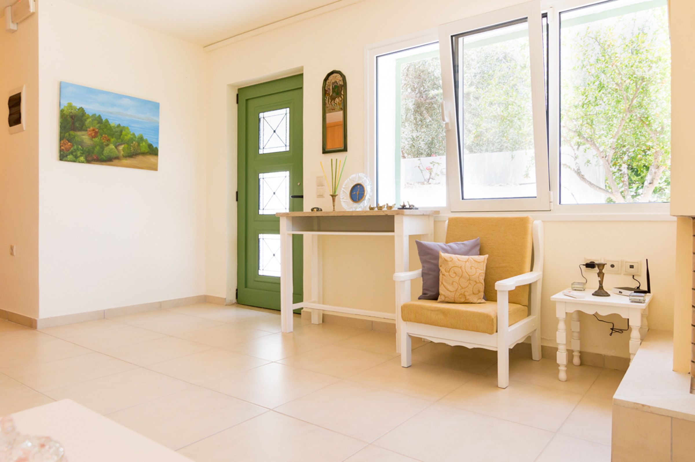 Una stanza luminosa con una porta verde, una sedia bianca con cuscini, un tavolino con una lampada e un quadro appeso al muro.
