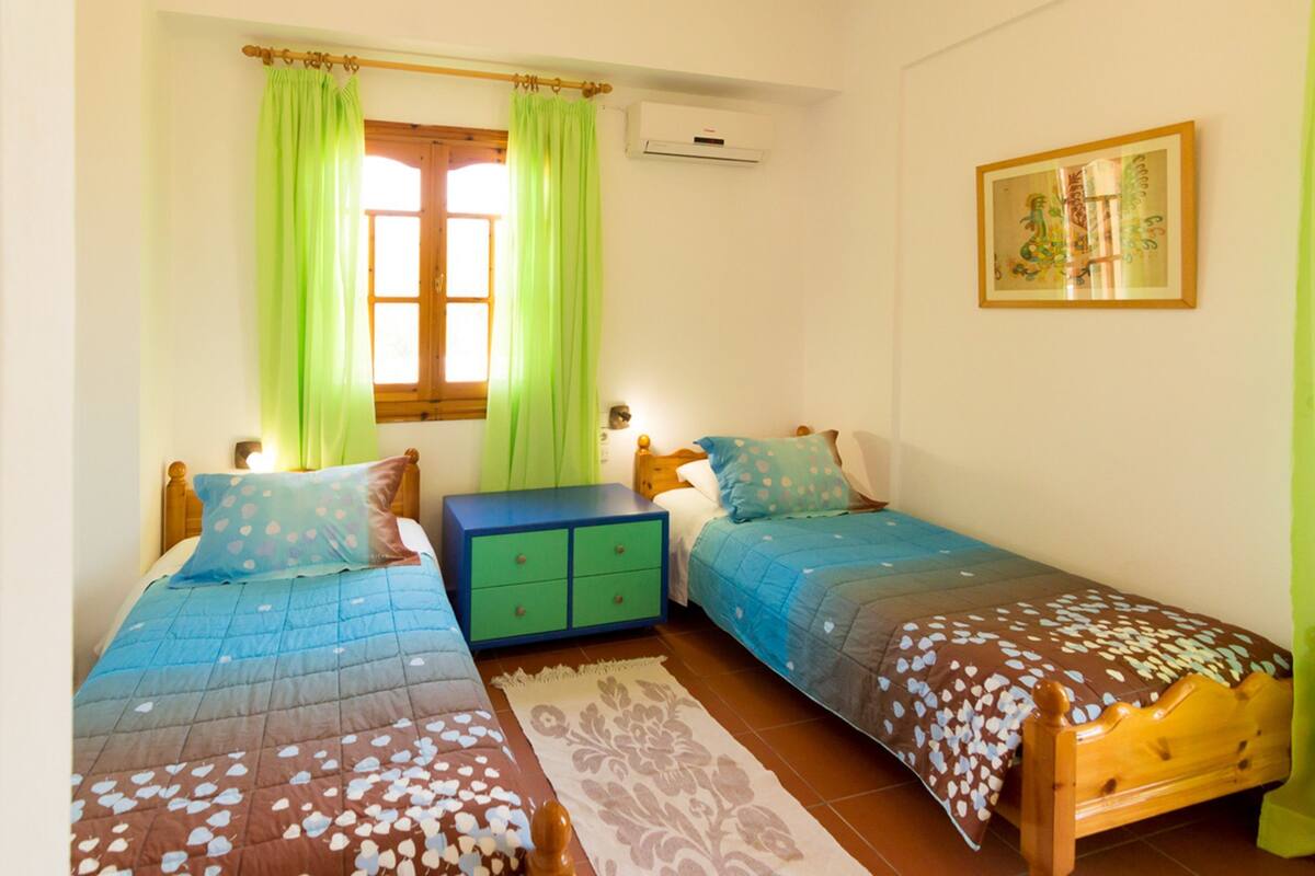 Een slaapkamer met twee eenpersoonsbedden, een groen nachtkastje, een houten dressoir en een raam met groene gordijnen.