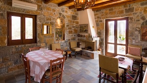 Une pièce aux murs de pierre, dotée d’une cheminée, de poutres en bois au plafond et d’un coin repas avec une table et des chaises.