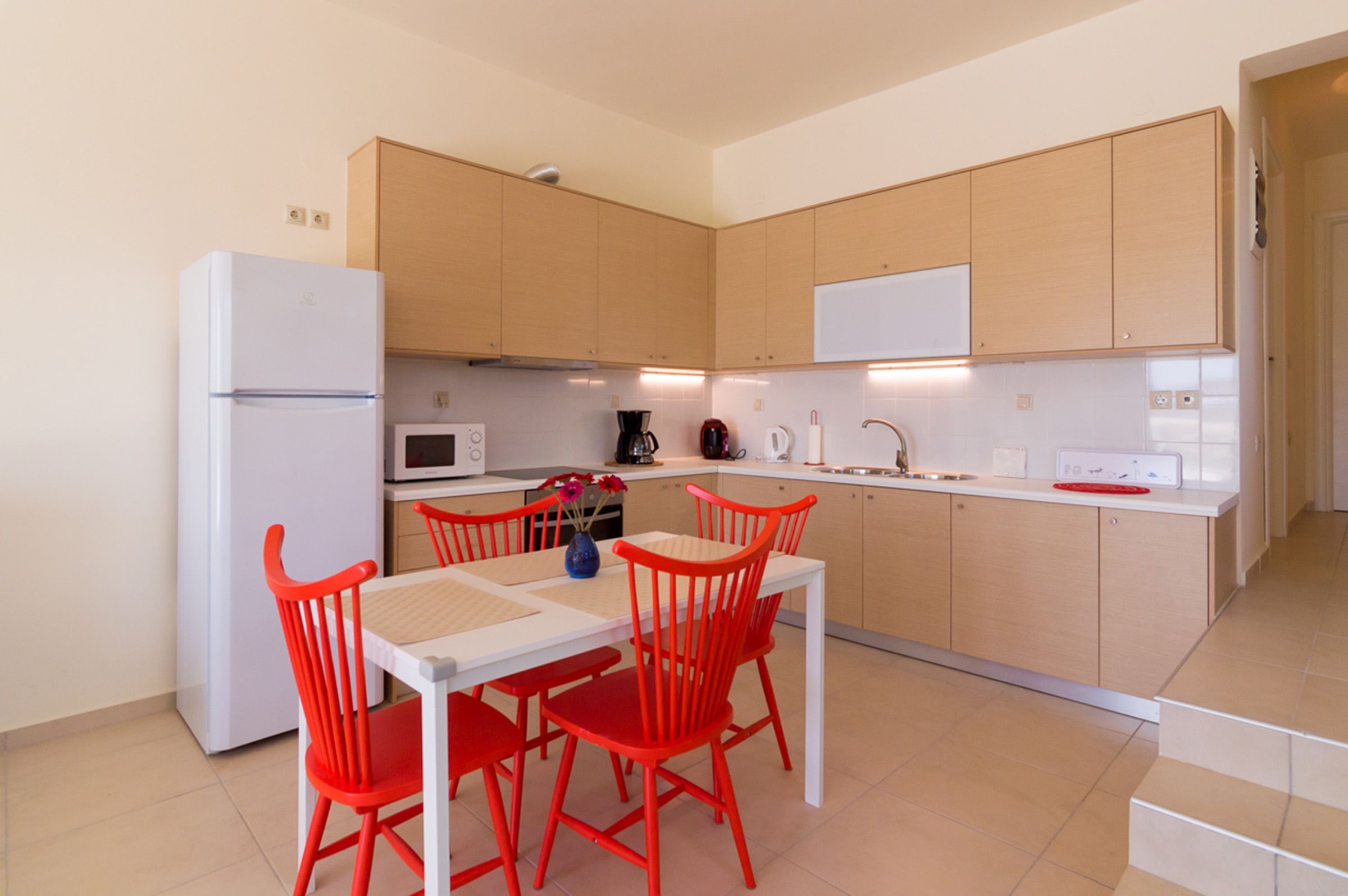 A compact kitchen with a refrigerator, microwave, coffee maker, and a dining table with red chairs.