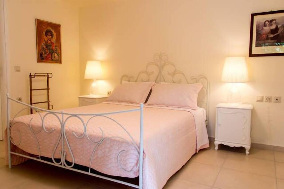 Een net opgemaakt bed met een metalen frame, twee nachtkastjes met lampen en een aan de muur hangend schilderij.