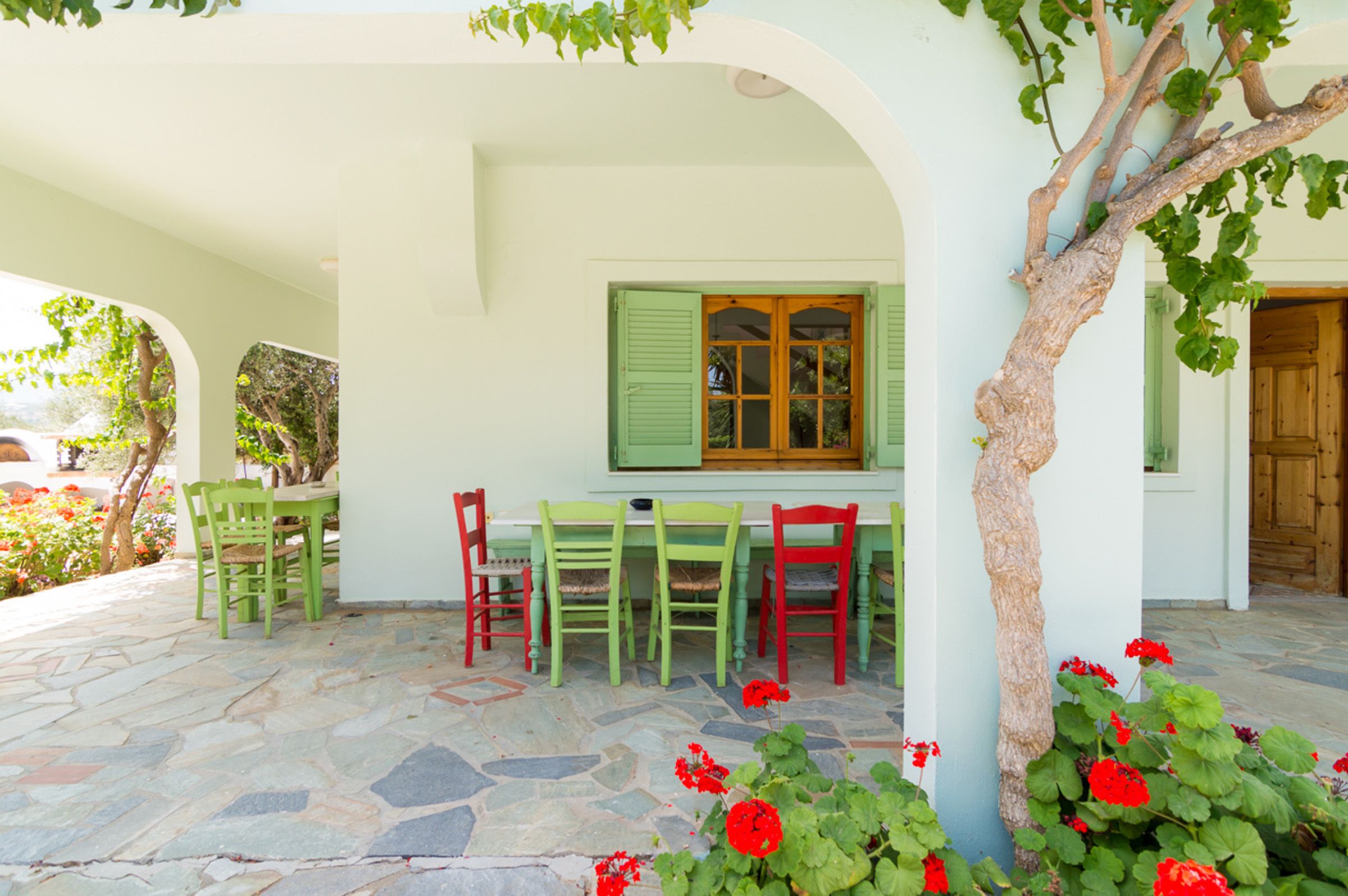 A covered outdoor seating area with a stone floor, green and red chairs, and a table.