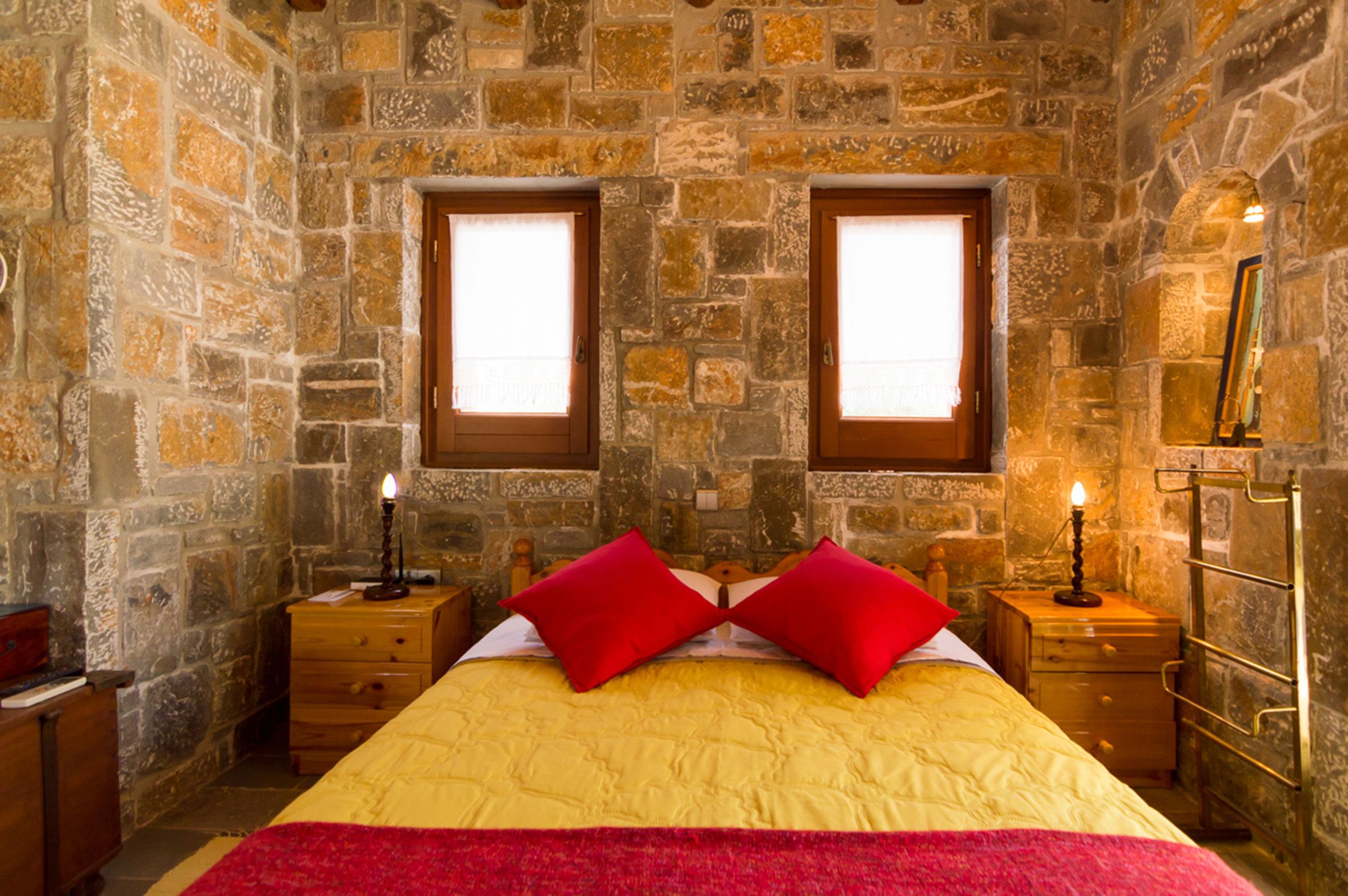 A room with a stone wall, a bed with red pillows, two bedside tables with candles, and a mirror on the wall.