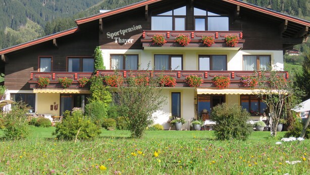 Un immeuble de deux étages doté de balcons ornés de jardinières, entouré d’un jardin luxuriant et surplombé par un paysage montagneux.