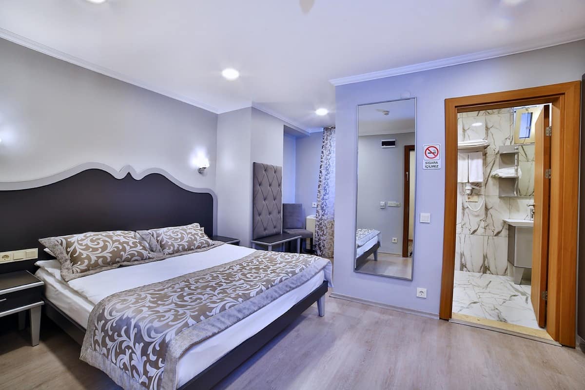 A modern bedroom with a large bed, bedside tables, a mirror, and a bathroom visible through an open door.