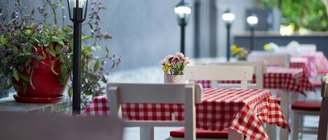 Un área de comedor al aire libre con mesas y sillas a cuadros rojos y blancos, plantas en macetas y farolas.