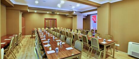 Un salón de conferencias con largas mesas y sillas, equipado con micrófonos y documentos, en una sala con paredes color beige y suelo de madera.