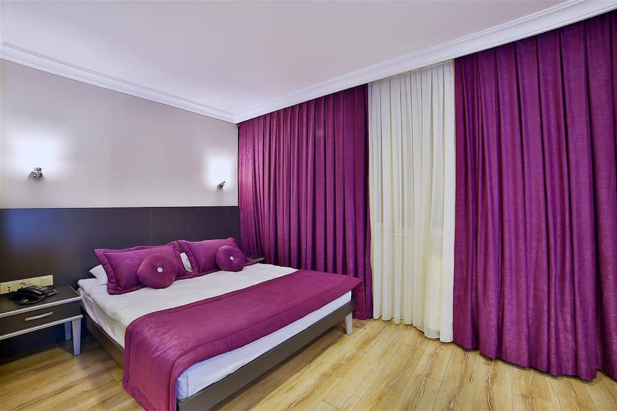 A modern hotel room with a large bed, purple and white curtains, a wooden floor, and a nightstand with a phone.