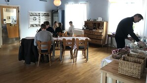 A dining area with wooden tables and chairs, a buffet table with food items, and a person serving food. - Helsingegården (Jarvso)