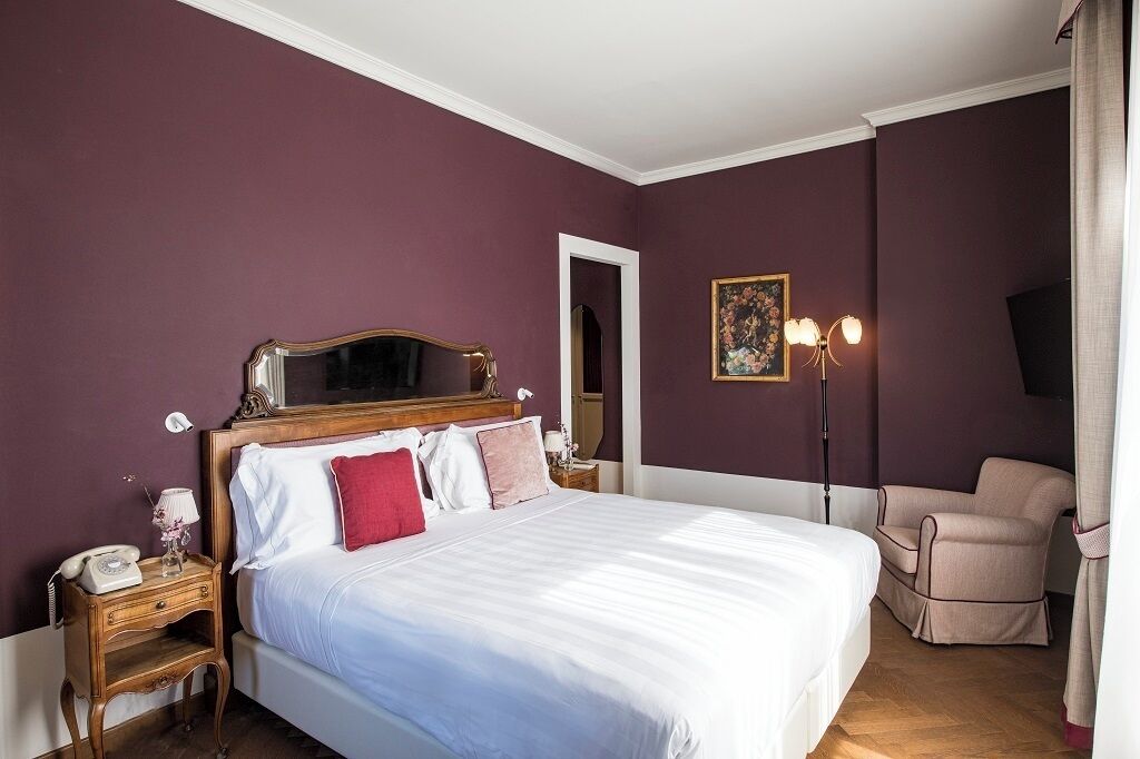Quarto com cama grande, mesa de cabeceira com abajur, cadeira e um quadro na parede.
