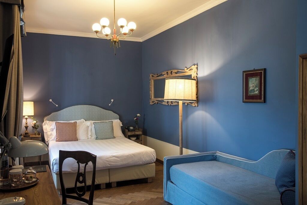A bedroom with a bed, a desk, a chair, a blue sofa, a chandelier, a mirror, and a framed picture on the wall.