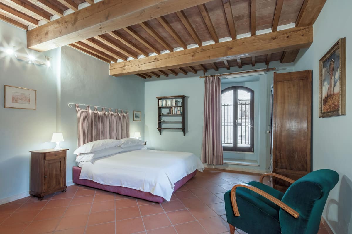A bedroom with a bed, a nightstand, a chair, a window with curtains, a wooden door, and a bookshelf.