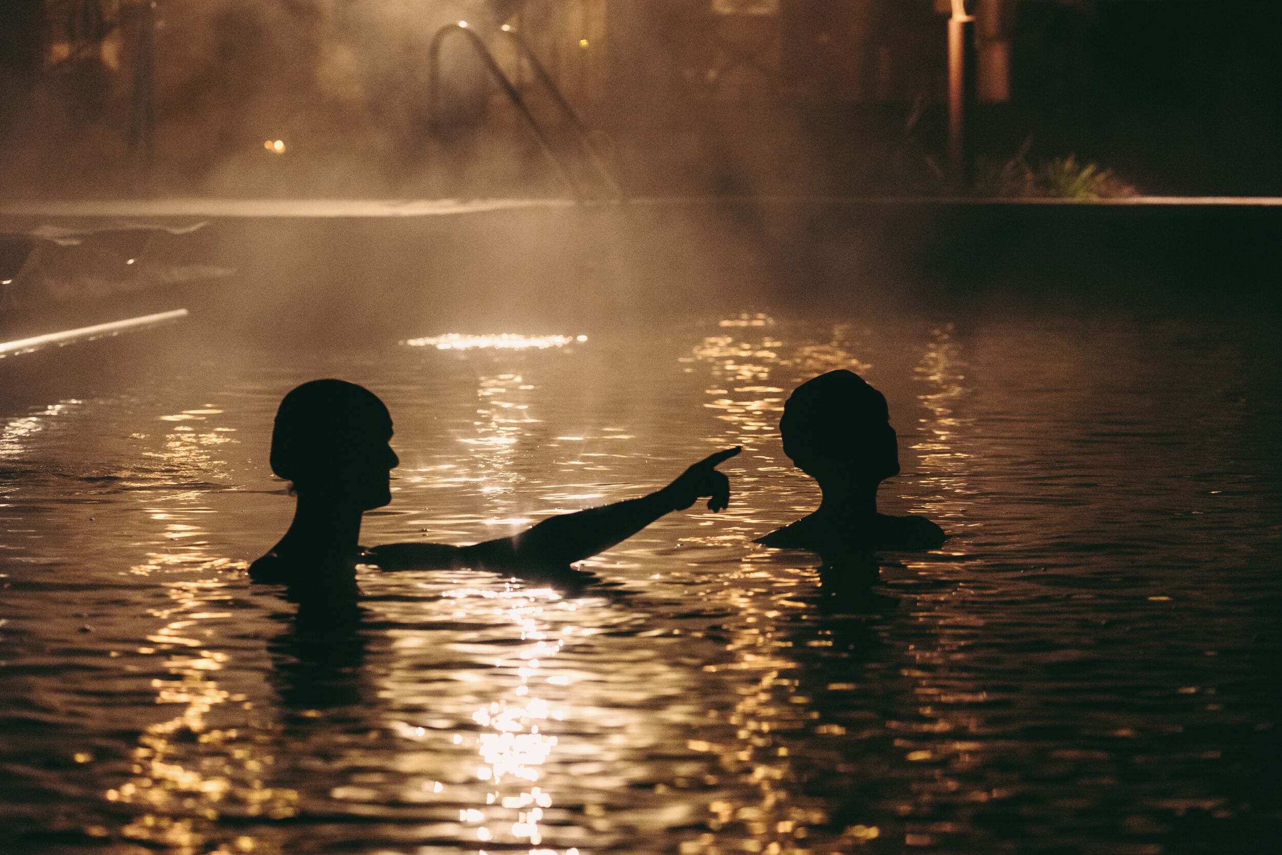 Deux personnes se tiennent dans un plan d’eau aux reflets changeants, peut-être une piscine, avec un arrière-plan brumeux et des lumières lointaines.