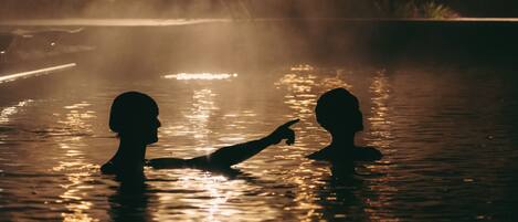 Deux personnes se tiennent dans un plan d’eau aux reflets changeants, peut-être une piscine, avec un arrière-plan brumeux et des lumières lointaines.