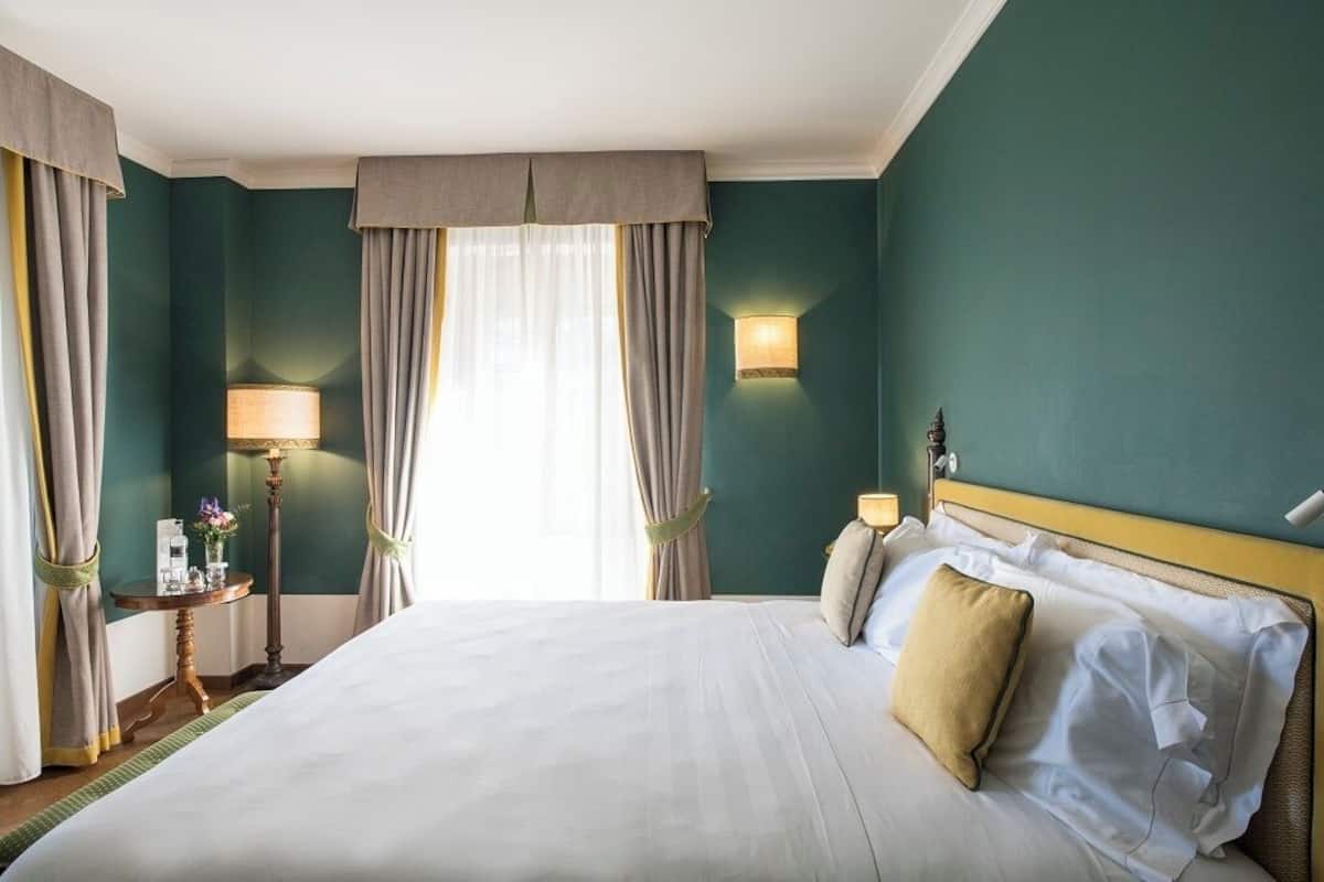 A hotel room with a large bed, a bedside table with a lamp, a wall-mounted light, and a view of the outdoors through the window.