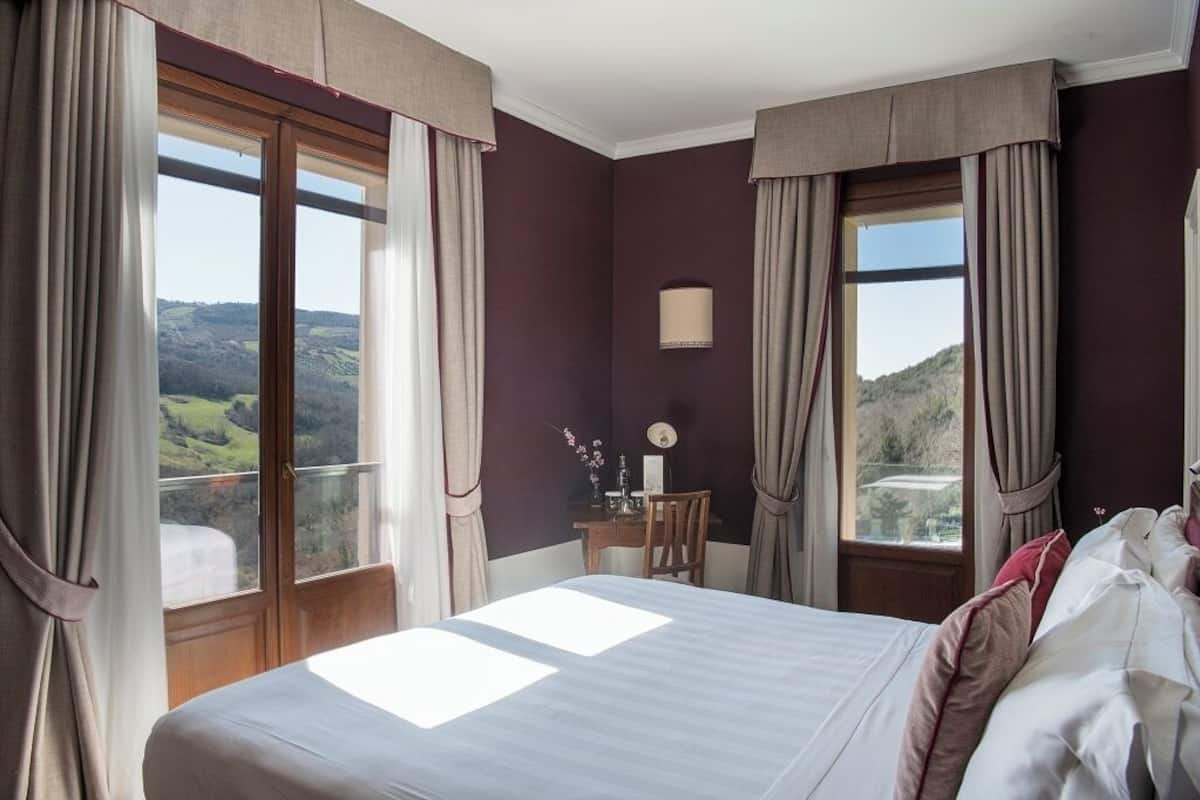 A bedroom with a large bed, two windows with curtains, a small desk with a lamp, and a view of the countryside.