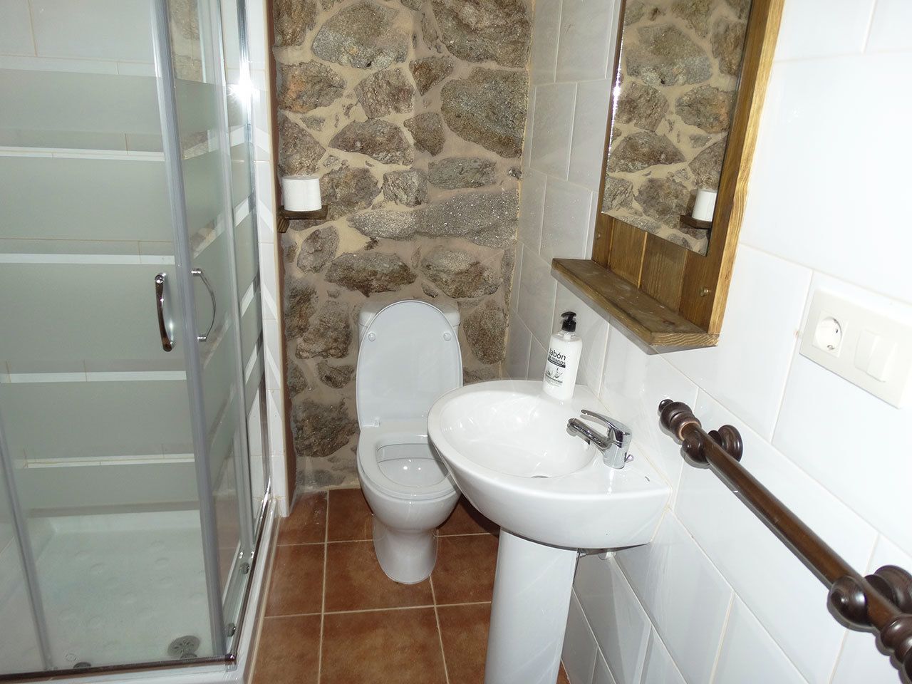 Un baño con cabina de ducha de vidrio, inodoro y lavamanos blancos, y una pared de piedra.
