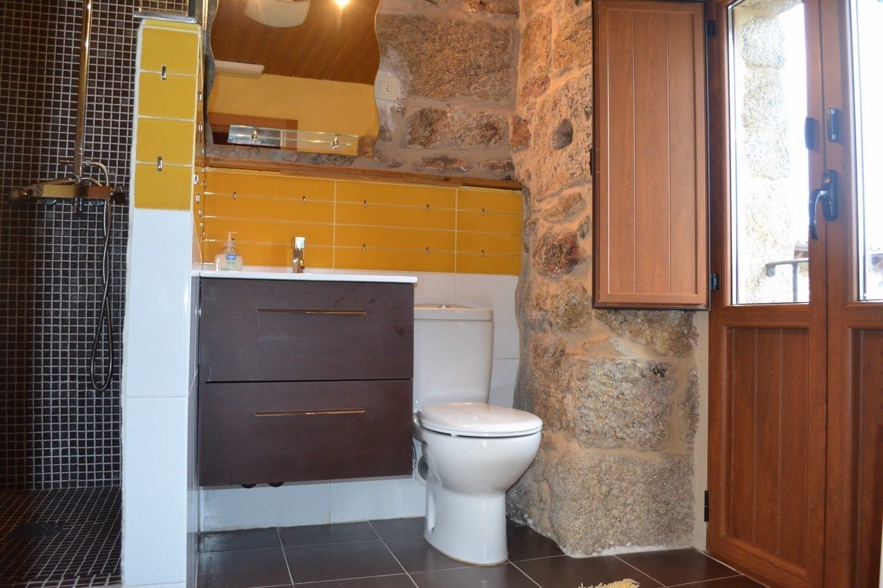 A bathroom with a stone wall, a wooden door, a white toilet, and a yellow-tiled vanity with drawers.