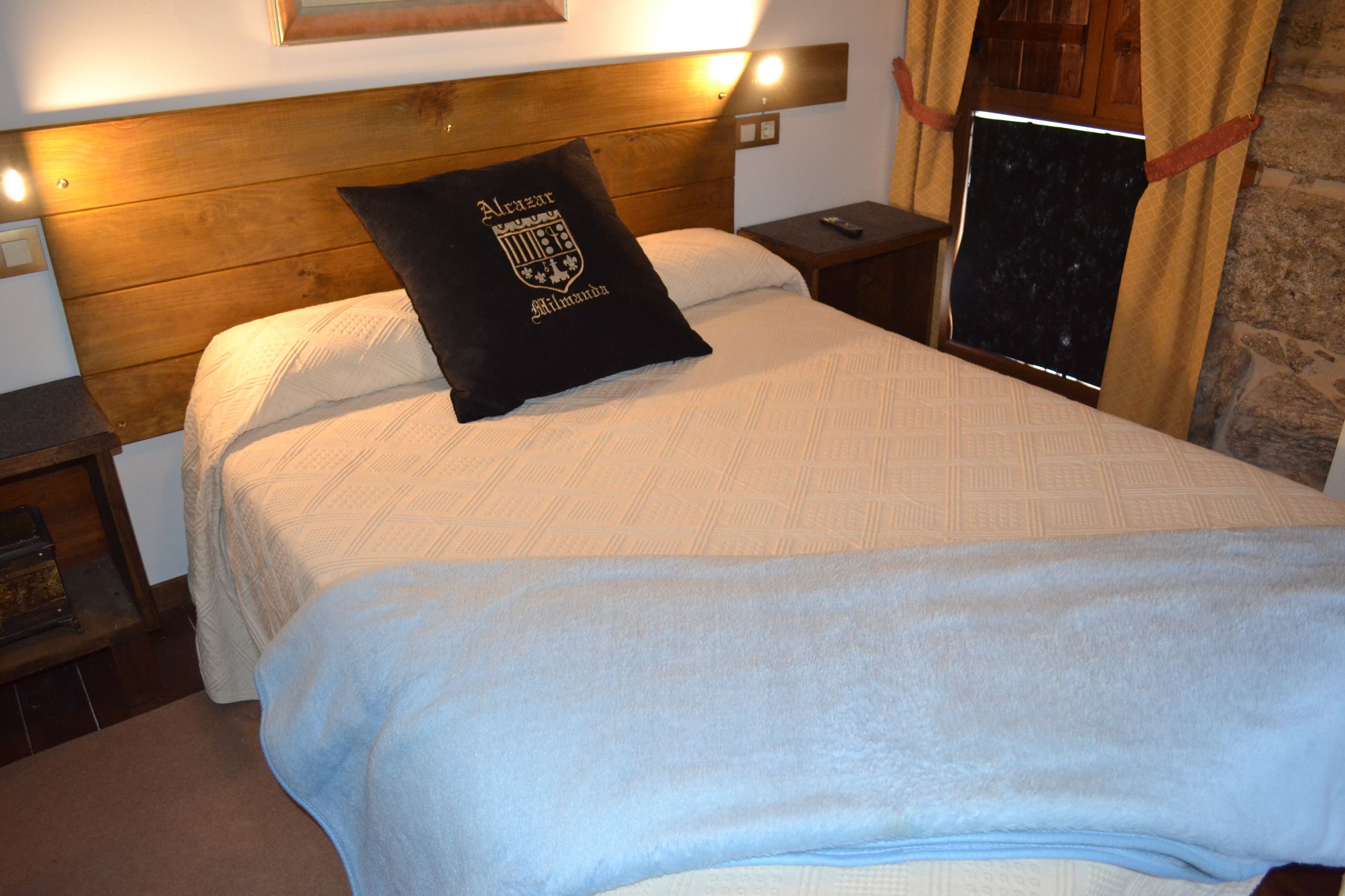 Una cama bien hecha con un almohadón negro que presenta un escudo, un cabecero de madera y una mesita de noche con un control remoto.