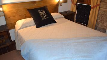 Una cama bien hecha con un almohadón negro que presenta un escudo, un cabecero de madera y una mesita de noche con un control remoto.
