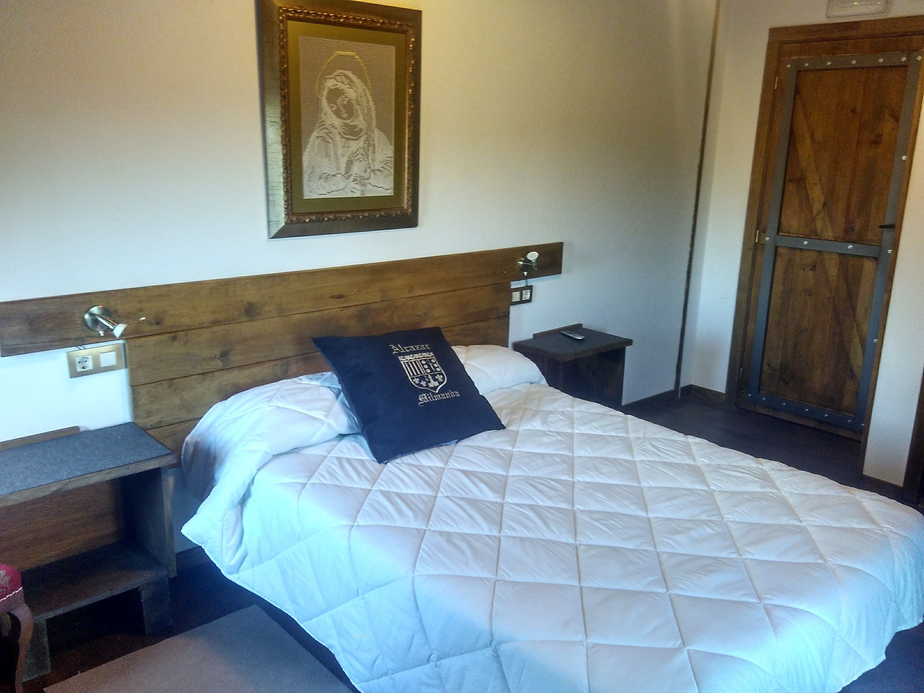 Una cama bien hecha con un edredón blanco acolchado, un almohadón con un escudo, una mesita de noche con una lámpara y un cabecero de madera.
