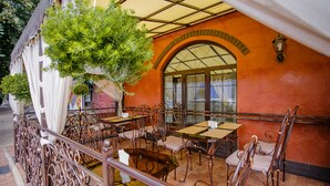 Un café al aire libre con sillas y mesas de metal, una planta colgante y un piso de baldosas.