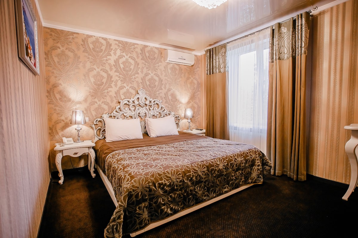 A hotel room with a large bed, ornate headboard, two bedside tables with lamps, and a window with curtains.