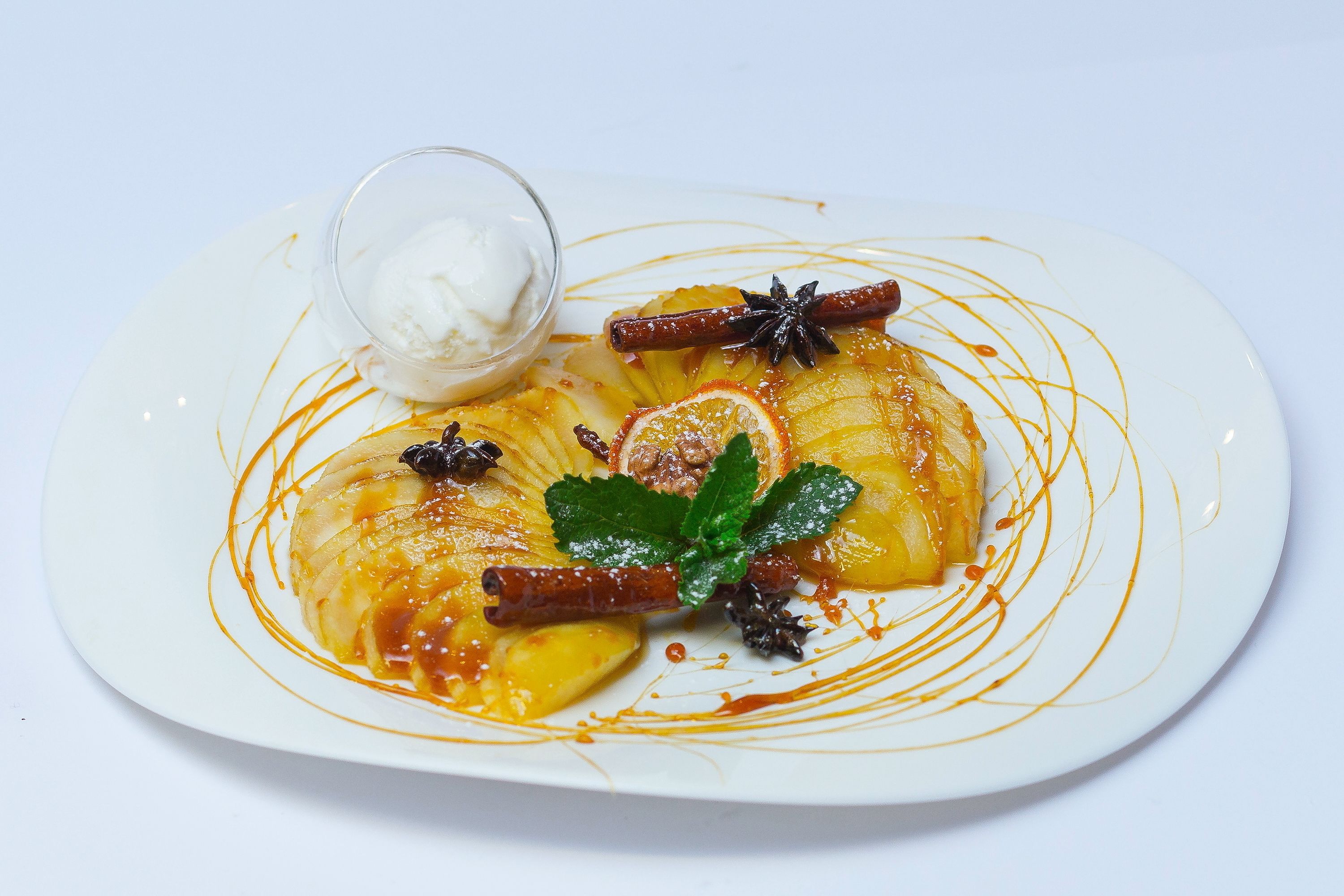 A dessert plate with a caramelized apple dish, garnished with star anise, cinnamon, and mint leaves, accompanied by a small bowl of whipped cream.
