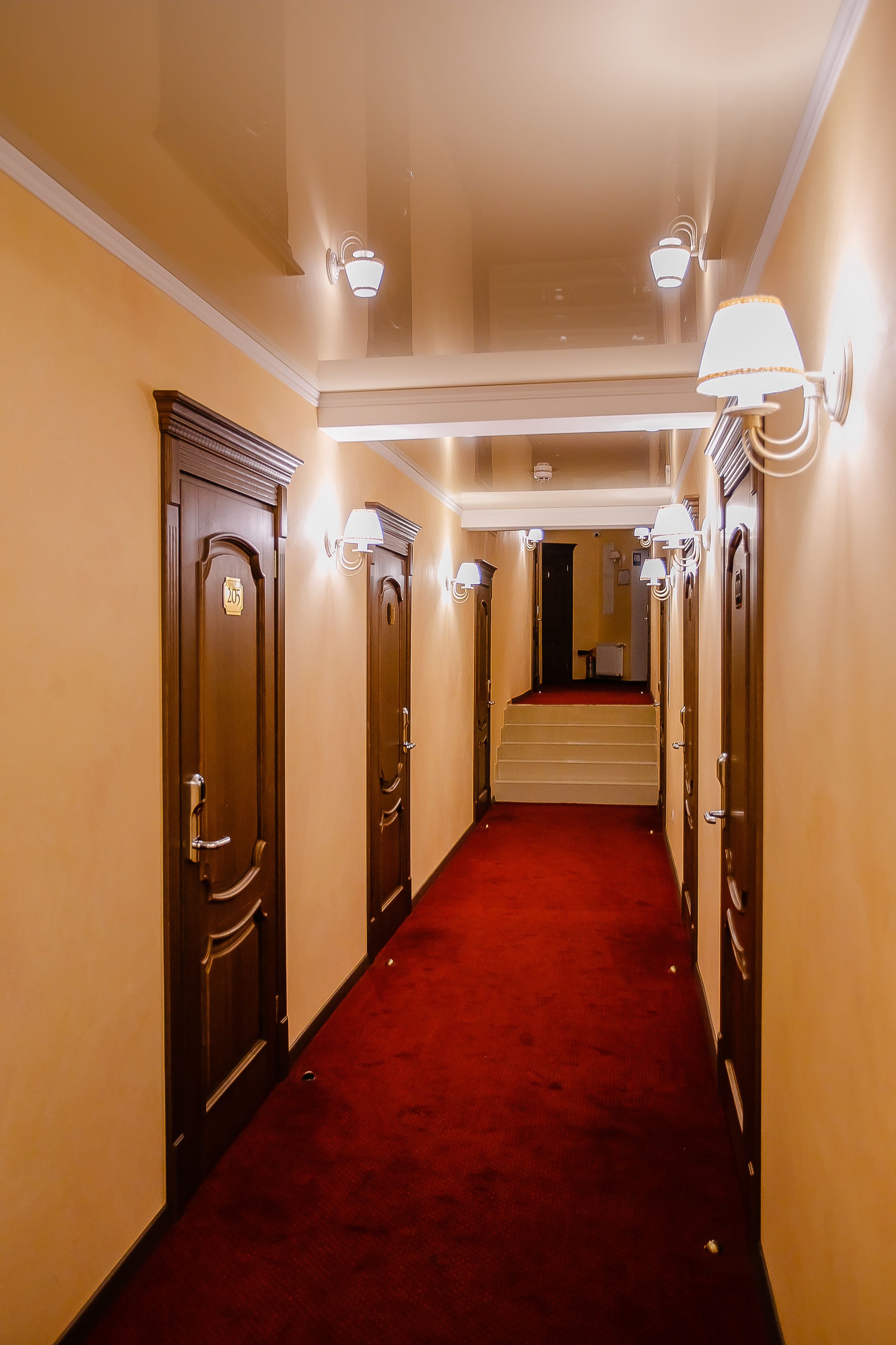 A hotel hallway with a red carpet, wooden doors, and wall-mounted lights.