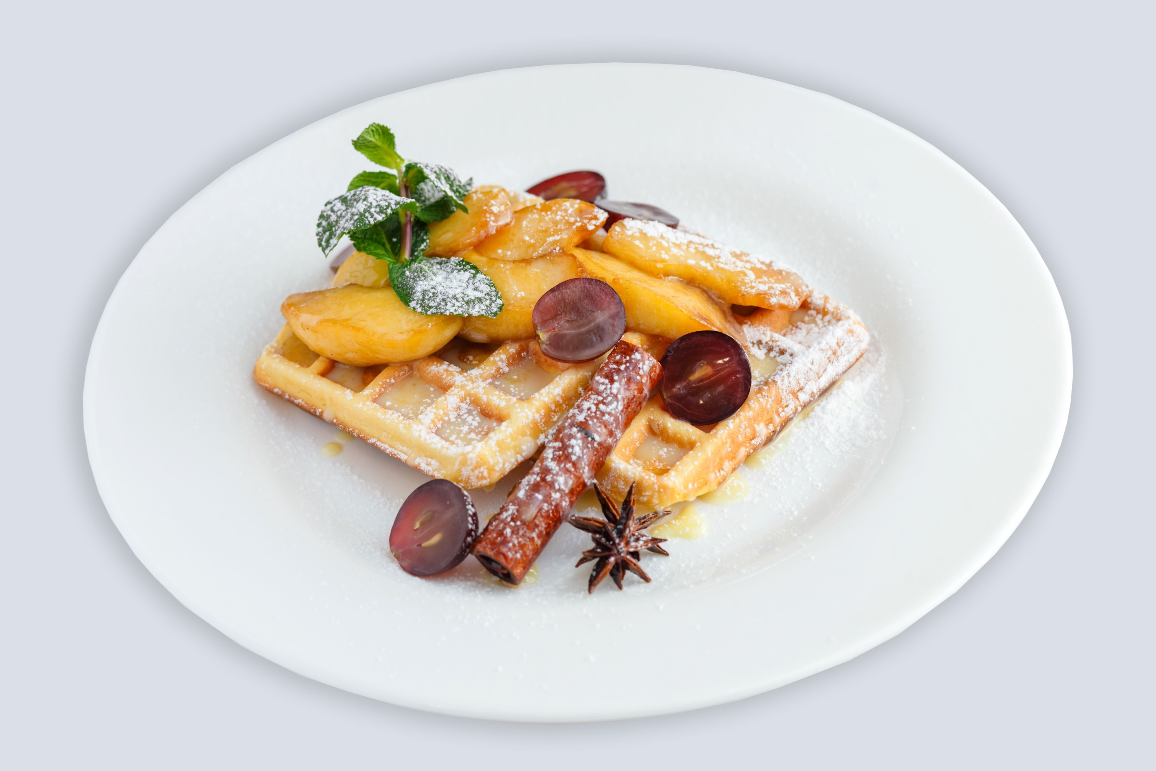 Une assiette de gaufres saupoudrées de sucre glace, garnie de tranches de prunes et d’une feuille de menthe.