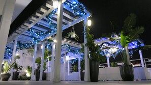 Un espacio al aire libre cubierto con columnas blancas, luces azules tipo guirnalda y plantas en macetas.