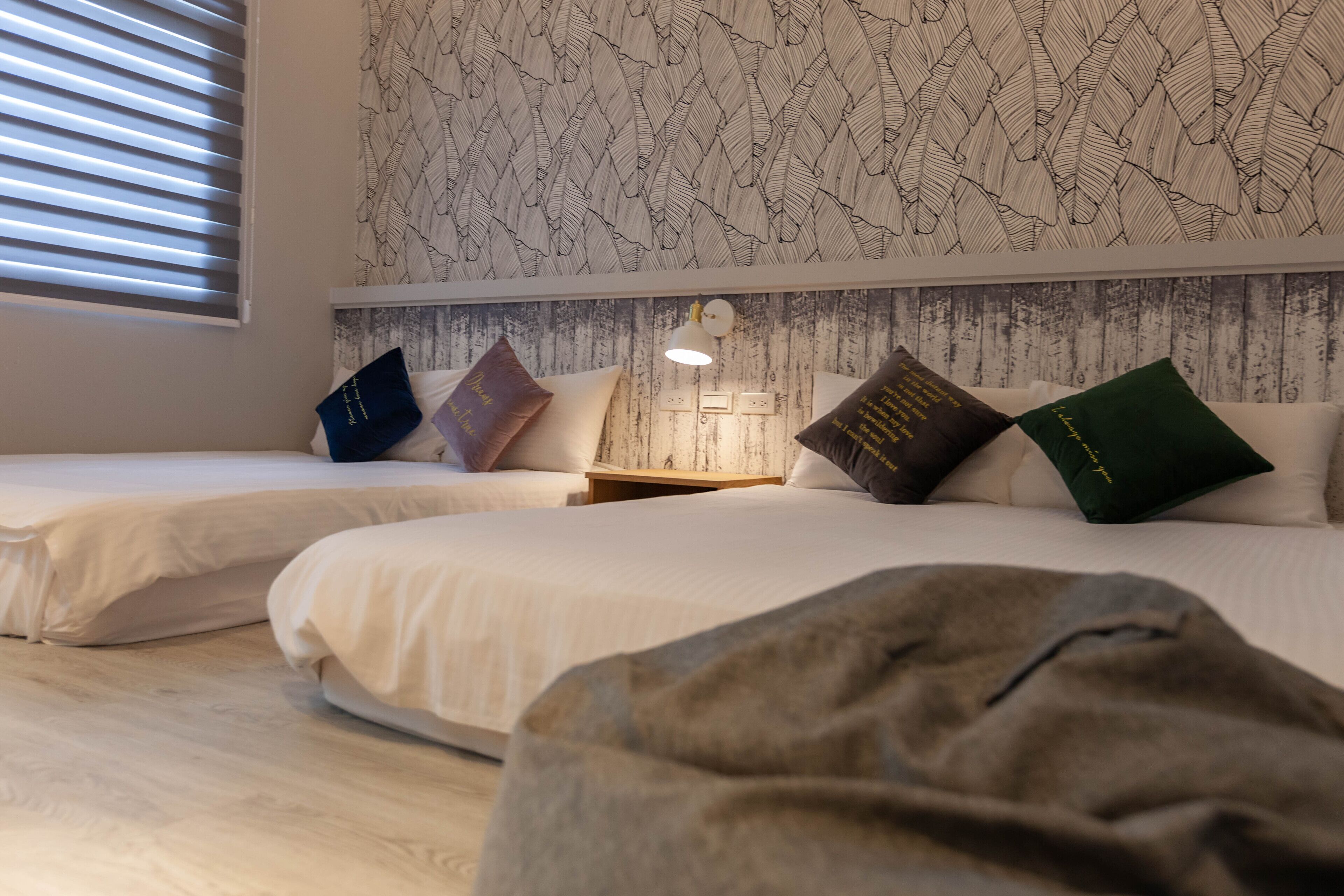 A bedroom with two beds, a patterned wallpaper, a nightstand with a lamp, and a window with blinds.