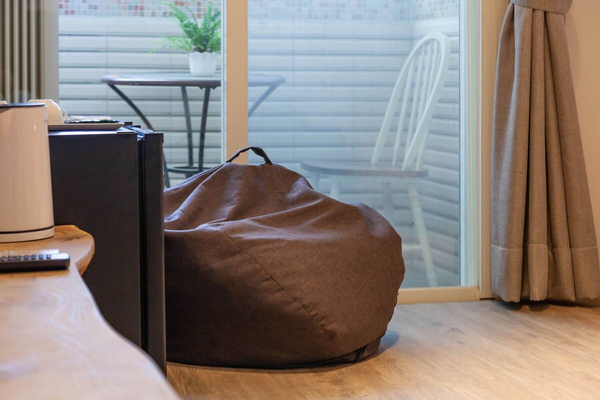 A room with a bean bag chair, a small table with a coffee maker, and a view of a balcony with a chair and a potted plant.