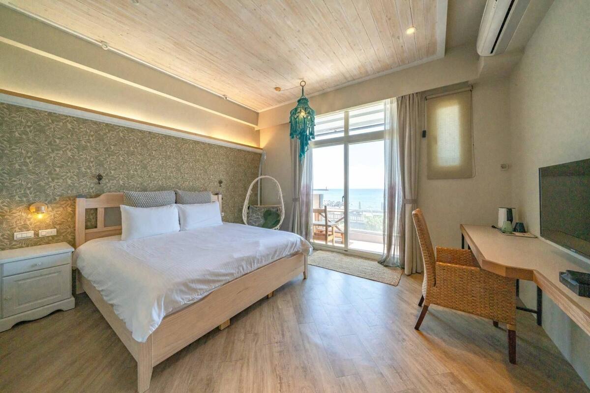 A bedroom with a bed, a desk, a chair, a TV, and a view of the sea.