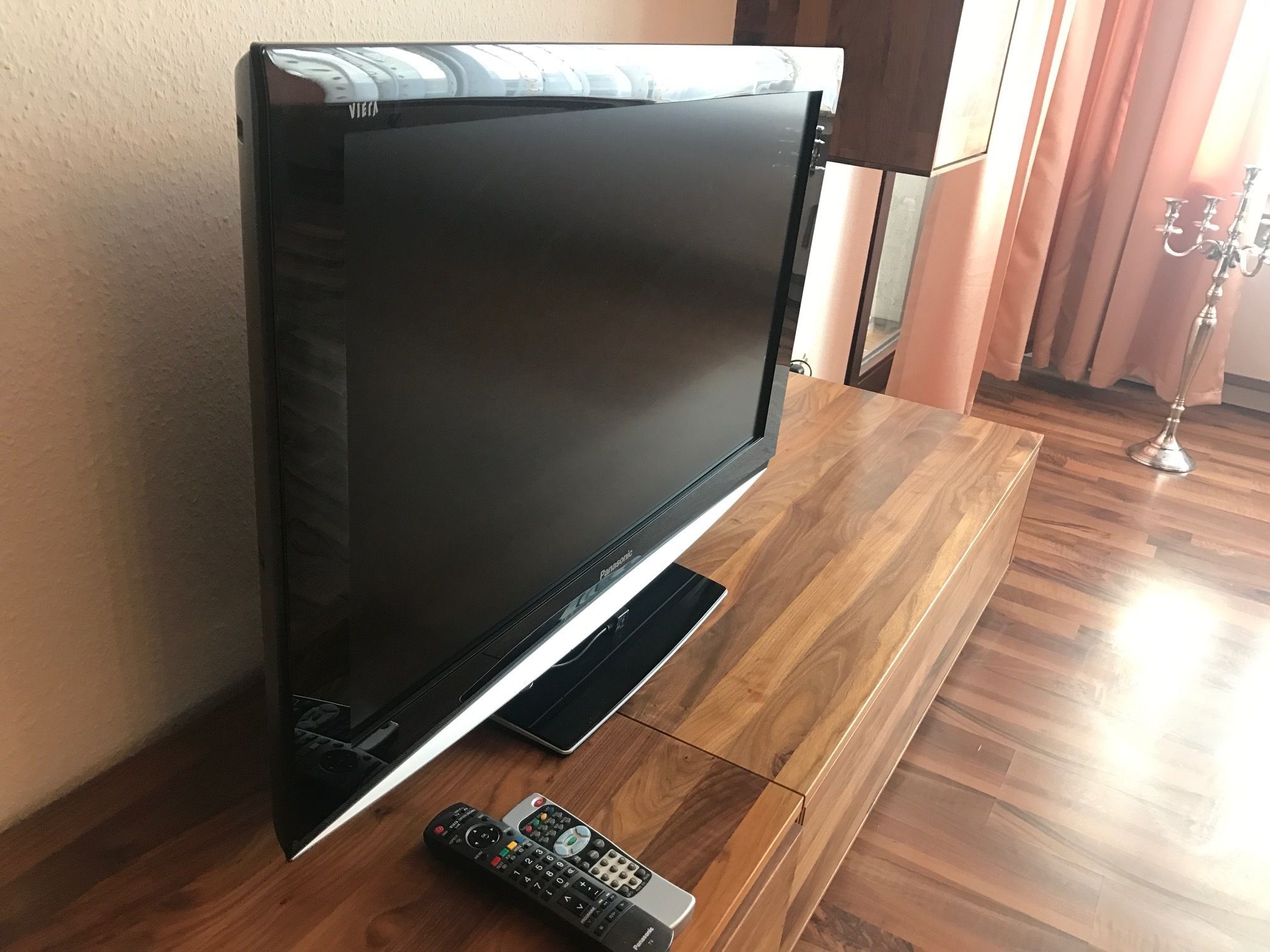 Una TV a schermo piatto su un supporto in legno in una stanza con una parete spoglia e una tenda.