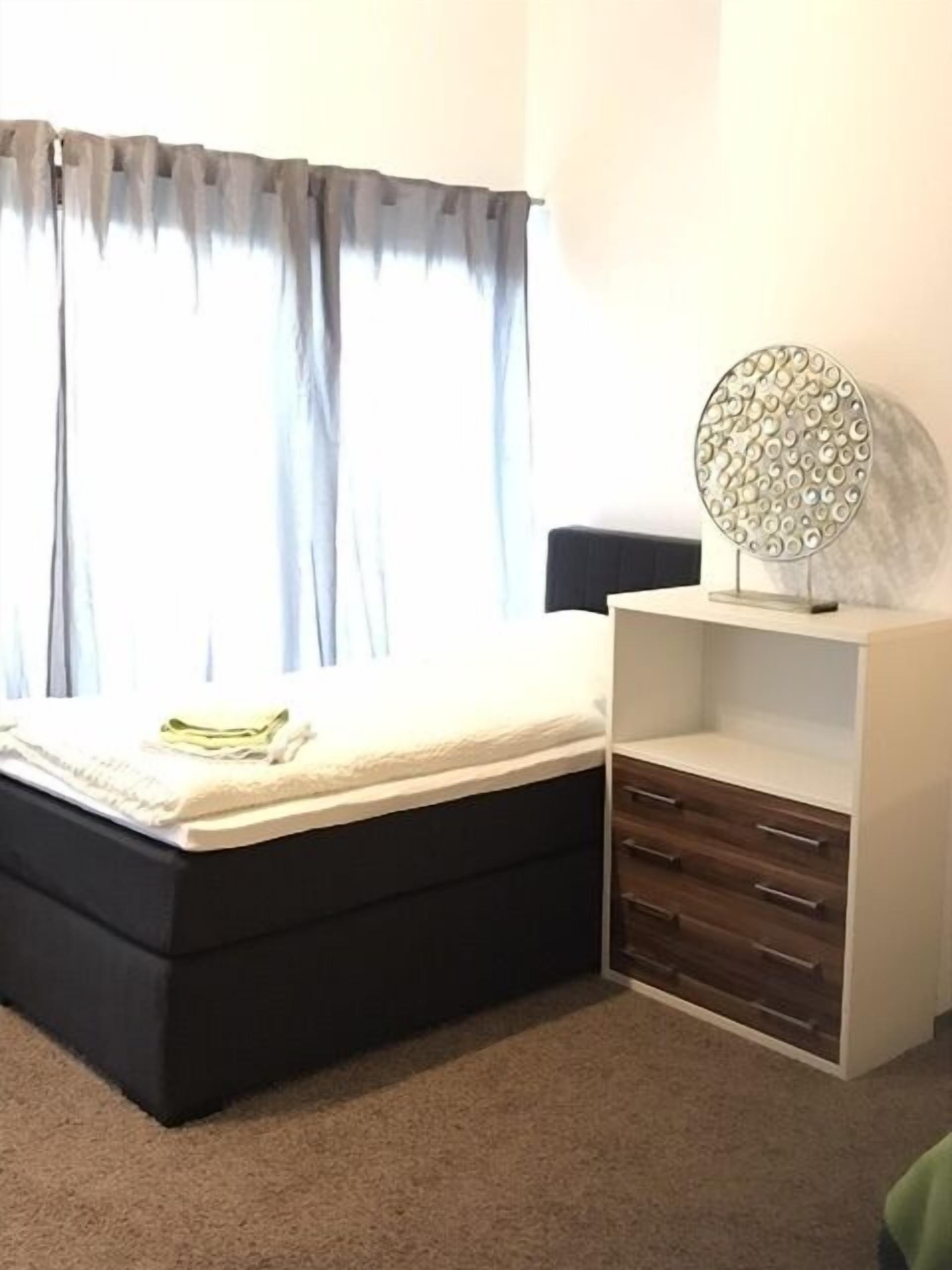 A bedroom with a bed, a bedside table, a wall-mounted shelf, and a window with curtains.