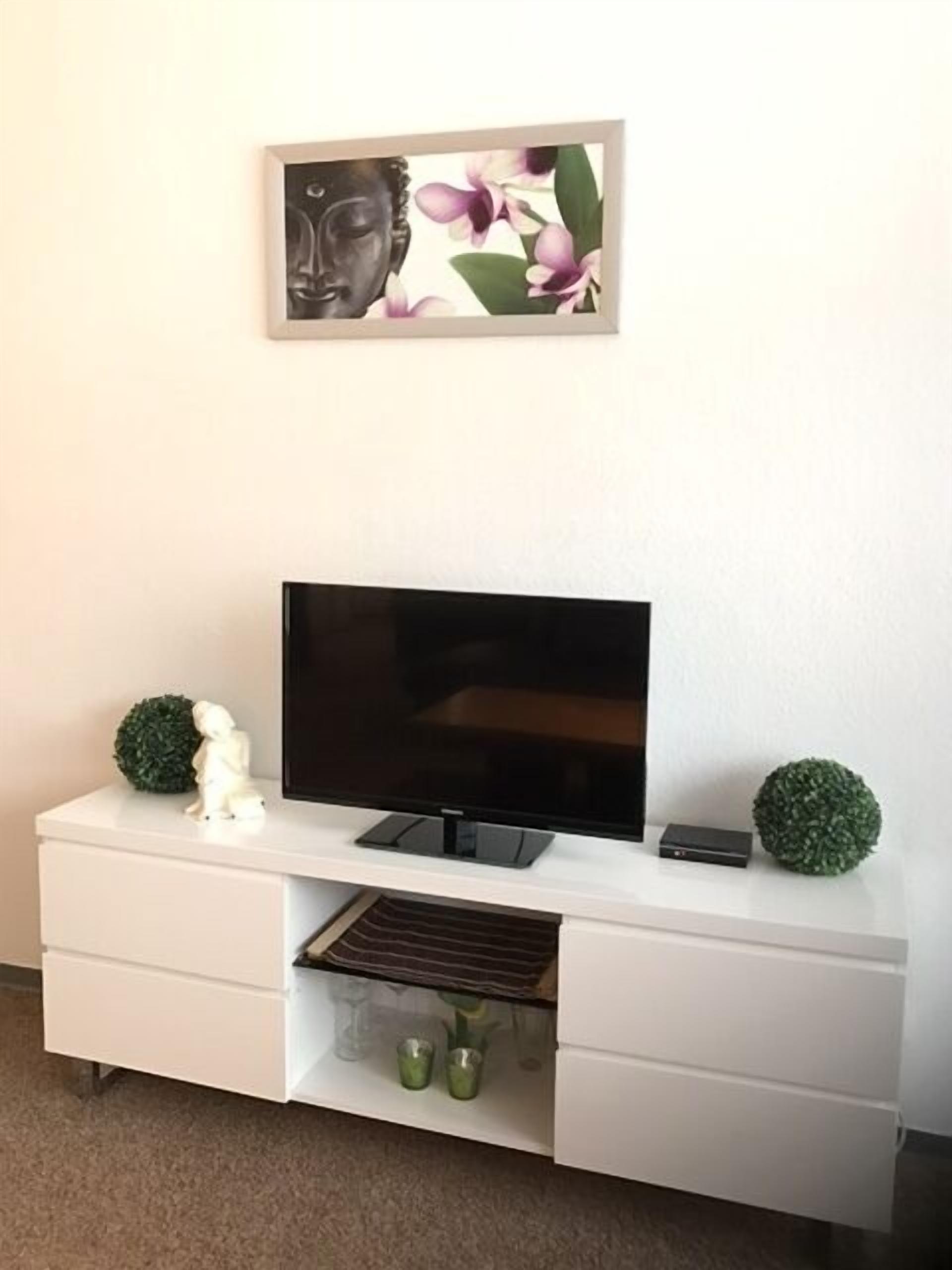 A modern living room with a flat-screen TV on a white entertainment unit, a decorative Buddha statue, and two green spherical ornaments.