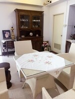 Una habitación con una mesa de comedor lista para cuatro personas, rodeada de sillas de mimbre. Se observa un aparador de madera con puertas de cristal al fondo, y una puerta a la derecha.
