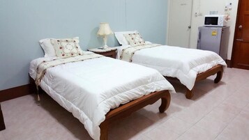 Dos camas individuales con estructura de madera y ropa de cama blanca, una mesita de noche con lámpara y un refrigerador al fondo.
