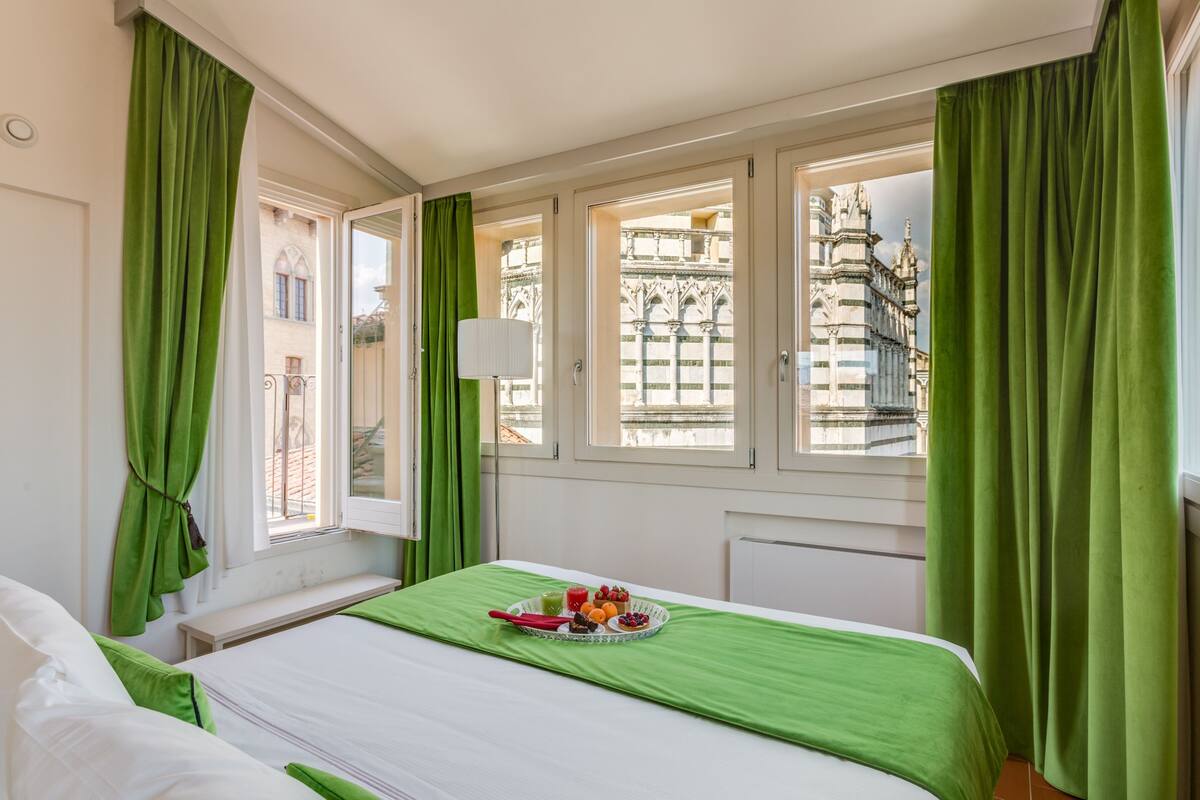 A hotel room with a bed, green curtains, a window with a view of a building, and a tray with fruit and juice on the bed.