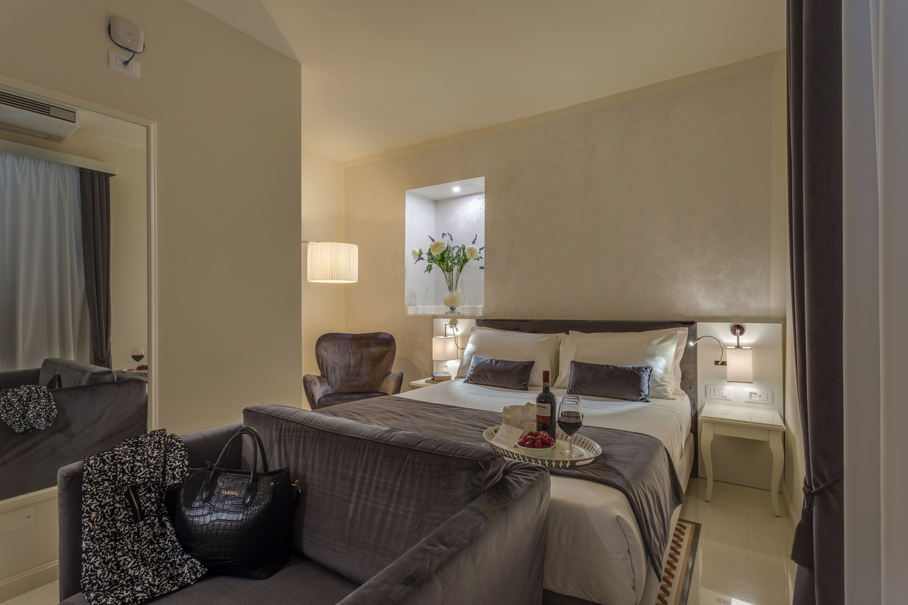 Quarto de hotel com cama, uma cadeira, mesa de cabeceira, abajur e bandeja com comida e bebida.