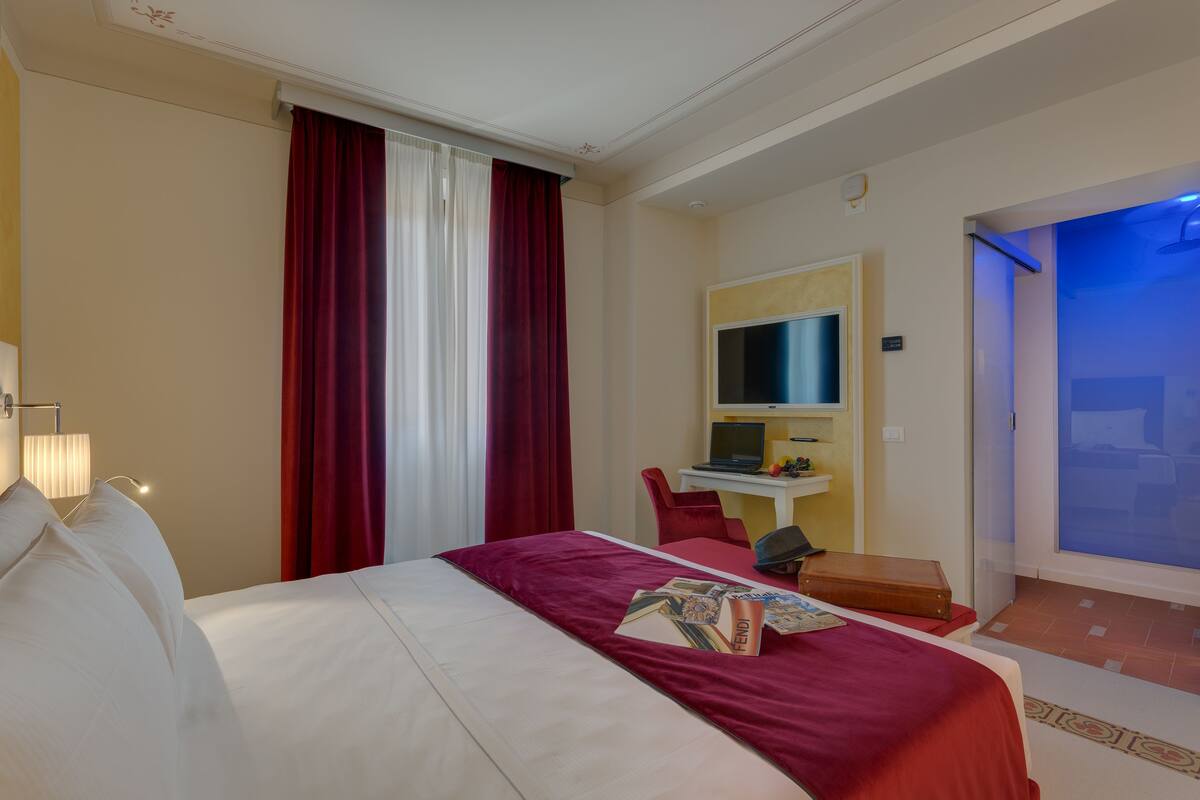 A hotel room with a bed, a desk, a chair, a television, and a bathroom with a blue-lit shower.
