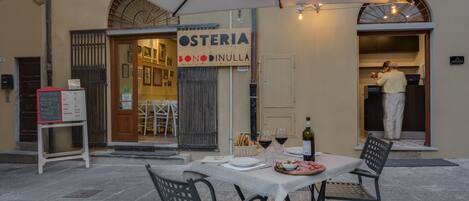 Un espacio de comedor al aire libre con una mesa para dos, un atril para el menú y un cartel que dice 'OSTERIA BONODINULLA'.