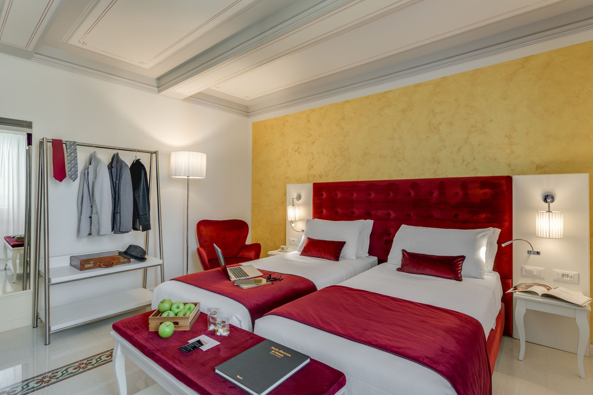 Ein Hotelzimmer mit zwei Betten, einem roten Kopfteil, einem Schreibtisch mit einem Laptop und einem Spiegel mit hängender Kleidung.