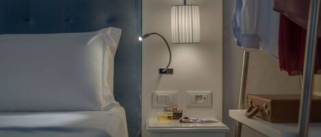 A hotel room with a bed, bedside table, lamp, and a small shelf with a suitcase.