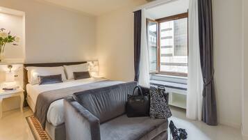 Habitación de hotel con cama, sofá, mesita de noche, lámpara, ventana con cortinas y vistas a edificios.