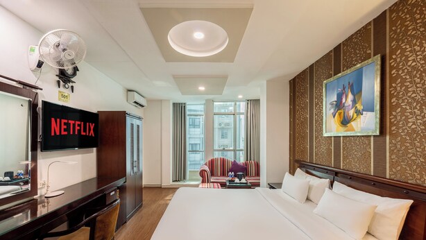 A hotel room with a large bed, a desk with a TV displaying "NETFLIX," a wall-mounted fan, and a painting. - A25 Premium Hotel - 20 Bui Thi Xuan (Ho Chi Minh City)